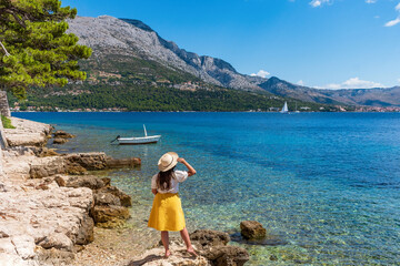 Back view of woman in stylish summer clothes standing on rocky beach in old Korcula town, Croatia