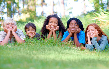 Nature, smile and portrait of children on grass in outdoor park, field or garden together. Happy, diversity and group of excited young kids relaxing and laying on lawn in woods or forest for summer.
