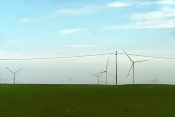 White wind turbines are on a green field, which also has a pole with an electricity wire. The sky is blue, with white clouds.