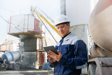 Engineer man in uniform with tablet computer controls loading of cement to mixer truck on concrete...