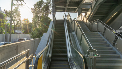 Photograph of an urban location, an escalator flanked by cement stairs. Handrails made of steel and glass, under roofs, public areas Outside the wall was a narrow alley, cars parked close together.