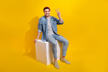 Fototapeta na wymiar Full length photo of friendly cheerful guy wear denim jacket sitting white platform showing v-sign isolated yellow color background