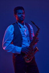 Naklejka premium Portrait of young man, solo performer, instrumental musician holds saxophone and looking at camera against dark background in neon light. Concept of jazz, blues, classy instrumental music, concert. Ad