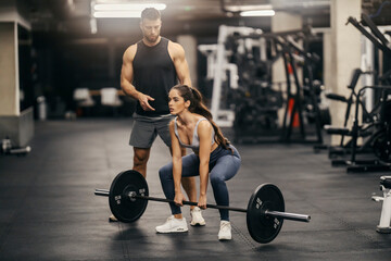 A strong sportswoman is doing deasdlifts in a gym while her trainer is helping her.