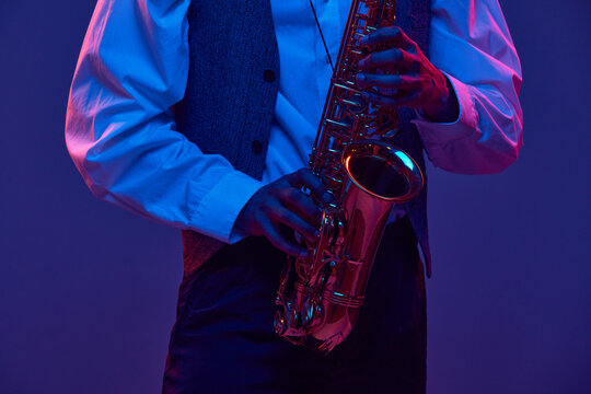 Cropped portrait of young man, saxophonist, musician in shirt and vest holds wind instrument against studio background in neon lighting. Concept of classical musical instrument, concerts, festivals.