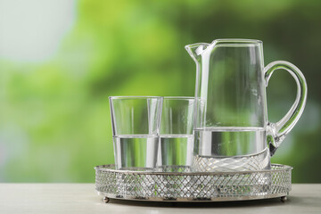 Jug and glasses with clear water on white table against blurred green background, closeup. Space...