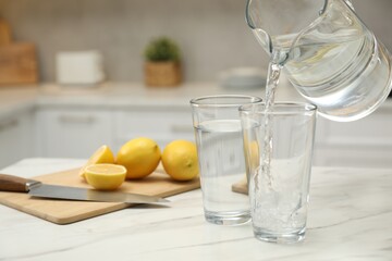 Pouring water from jug into glass on white table in kitchen, closeup