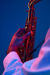Cropped portrait of young man holds sax and playing blues melodies against blue background in neon...