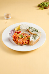 Salmon and avocado tartare with crispy bread and cream cheese, elegantly plated with lemon and dill on a white plate against a yellow background