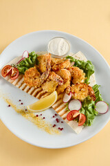 Tempura shrimp with tartar sauce on a bed of lettuce, presented on a white plate with lemon and tomato accents, against a soft yellow backdrop