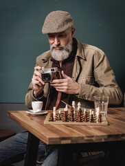 Portrait d'un homme photographe quinquagénaiore agé senior avec une barbe et un béret tenant un appareil photo argentique et assis à une table avec un jeux d'échec