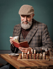 Portrait d'un homme agé avec une barbe qui lit un livre rouge en buvant un café assis à un bar en jouant aux échecs