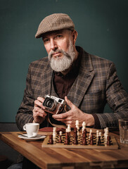 Portrait d'un homme photographe élégant en costume quinquagénaiore agé senior avec une barbe et un béret tenant un appareil photo argentique et assis à une table avec un jeux d'échec