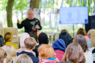 Focused audience listening to a presenter at an outdoor educational workshop in a park setting.