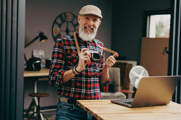 Portrait d'un homme photographe hipster situé dans un studio atelier et qui tient un appareil photo vintage en souriant devant un ordinateur