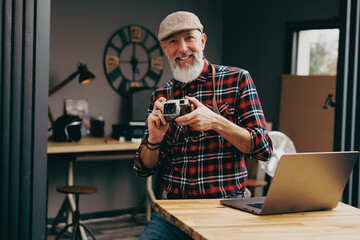 Portrait d'un homme photographe hipster situé dans un studio atelier et qui tient un appareil photo vintage en souriant devant un ordinateur - 724588033