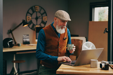 Hipster assis souriant quinquagénaire senior élégant et stylé qui travaille sur un ordinateur dans un atelier créatif vintage et qui porte un gilet et une casquette et qui bois un café