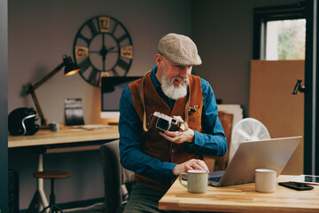 Homme photographe assis souriant quinquagénaire senior hipster élégant et stylé qui travaille sur un ordinateur dans un atelier créatif vintage et qui tient un appareil photo en buvant un café - obrazy, fototapety, plakaty