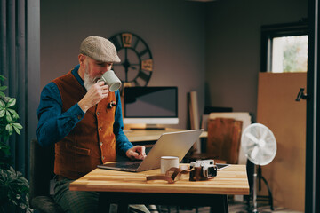 Homme photographe hipster élégant et stylé qui travaille sur un ordinateur dans un atelier créatif vintage et qui porte un gilet et un béret  en buvant un café