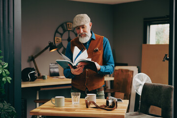 Portrait d'un homme quinquagénaire senior hipster élégant et stylé qui tient un livre pour le lire dans un atelier