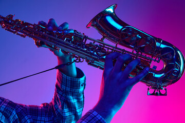 Cropped portrait of jazzman playing saxophone against gradient blue-pink background in neon light....