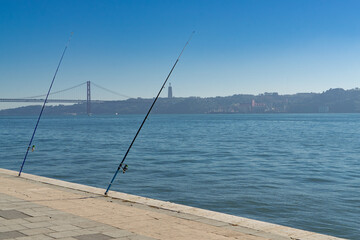 Two fishing rods in Lisbon in Portugal, the bridge of the 15th April and the Cristo Rei in the background - 724605254