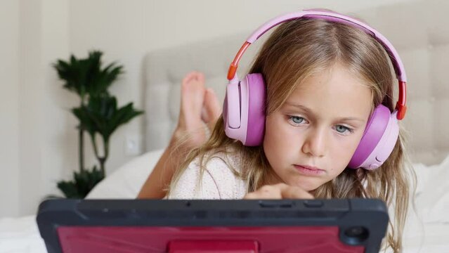 Cute little kid girl using digital tablet technology device lying on bed at home. Small child surfing Internet play game read book in online library. Children tech addiction