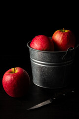 Top view of metal bucket with red apples on wooden table with apple and knife, black background, vertical, with copy space
