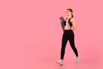 Active woman carrying yoga mat and water bottle walking on pink background