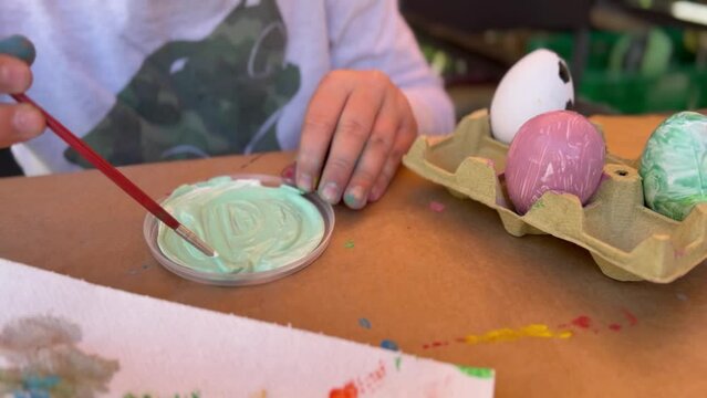 11 year old boy mixing paints with a brush to paint Easter eggs sitting on a table in a yard. Kid's handicraft. Sunny outdoor.