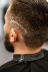 Obraz premium A young man with a beard in a black cape sits on a chair in a barbershop. The client is waiting for a haircut of the beard.