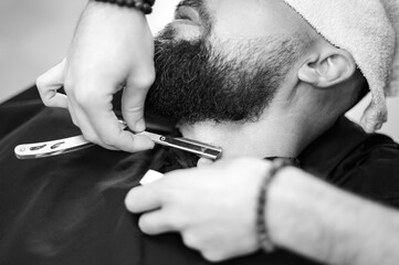 A barber shaves the neck of a bearded customer with dangerous razor. Shaving the contour of the beard for the correct shape.