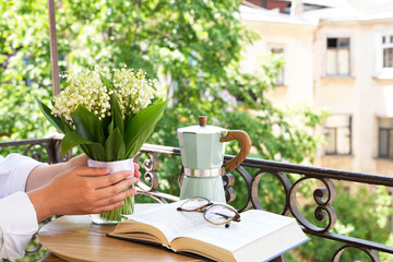 Woman hands with bouquet of flowers in vase on the balcony with book, glasses and coffee pot . Female holding bouquet of lilies of the valley. Spring holidays, Easter, mother's day.