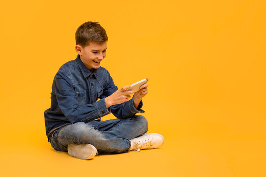 Cheerful teen boy playing games on smartphone while sitting on yellow background