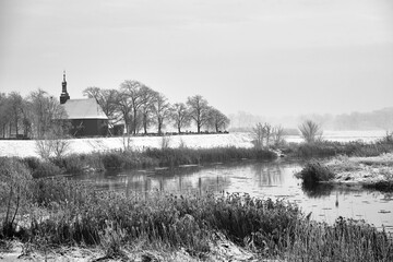 wooden church over the Warta river and snow-covered bushes and trees during winter - obrazy, fototapety, plakaty
