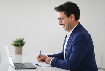 Cheerful caucasian adult businessman in suit at table with laptop, make notes