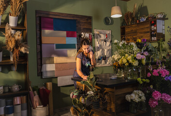 Young woman working in her flower shop talking on the phone and receiving an order for a bouquet of...