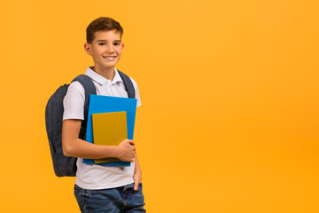 Smiling schoolboy carrying backpack and textbooks standing against yellow studio background