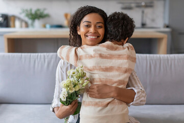 Smiling black mother receiving hug from son and holding bouquet of flowers