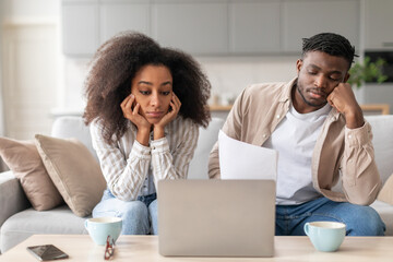 Upset African American Couple Looking At Laptop Holding Documents Indoor