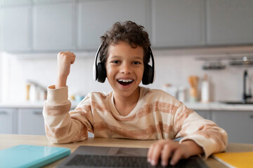 Cheerful black boy wearing wireless headphones using laptop computer and celebrating success