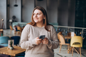 Young happy woman in glasses smiling and using smart phone in a cafe. Person using smartphone indoors. Online education, order, working or shopping concept