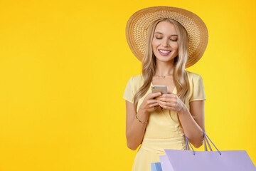 Young blonde woman in hat over isolated yellow background holding shopping bags and writing a message with her mobile phone for online purchasing