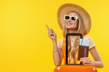 Young smiling woman pointing in vacation with suitcase and passport over isolated yellow background. Traveler on trip voyage tourist showing copy space - obrazy, fototapety, plakaty