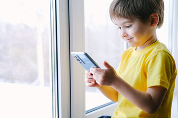 Little toddler boy is holding the smartphone at home. He is wearing bright yellow t-shirt. Playing video games like a social problem - obrazy, fototapety, plakaty