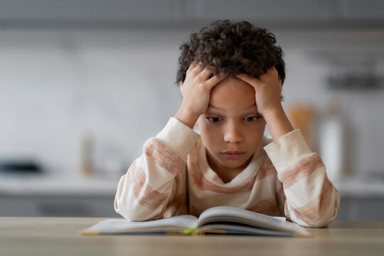 Stressed Little Black Boy Reading Book At Home And Touching Head