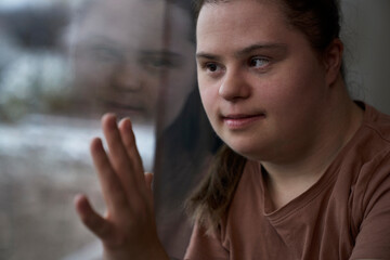 Pensive down syndrome woman standing next to window and looking away