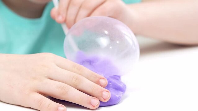 A child plays with a toy made of bright purple slime, inflates bubbles with a tube, a fashionable liquid toy sticks to his hands and fingers.