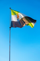 Huge flag of Extremadura fluttering in the wind on a flagpole against a blue sky.