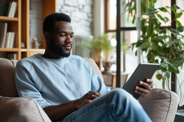 African American man relaxing at home he is sitting on the armchair and connecting with his digital tablet contemporary design interior